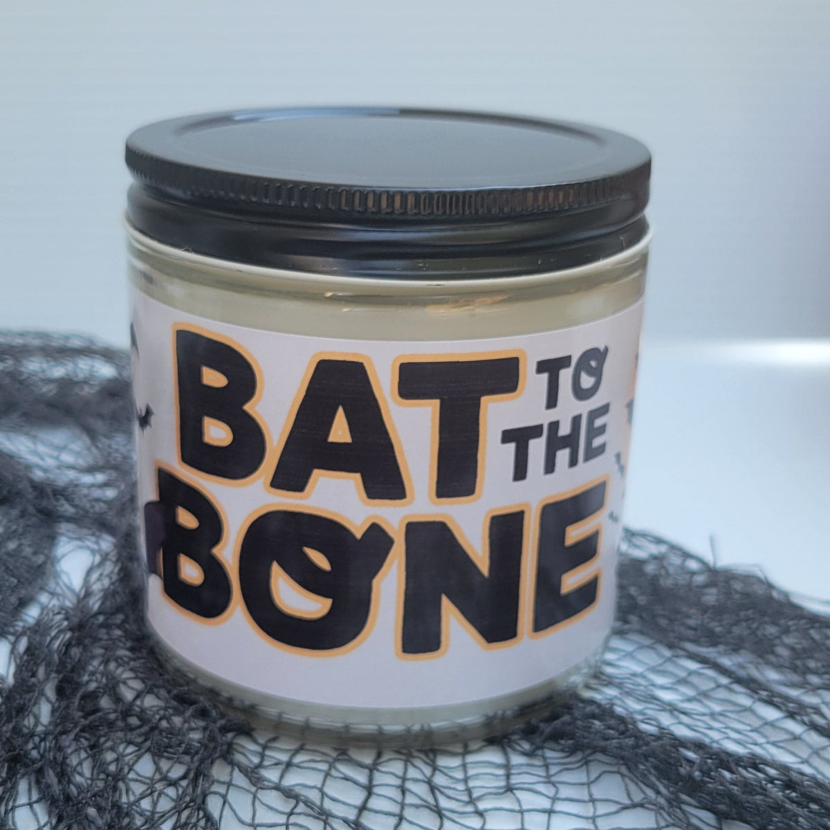A glass jar candle with a black lid and a white label reading 'BAT TO THE BONE' in black and orange lettering, styled on black spiderweb mesh against a light gray background.