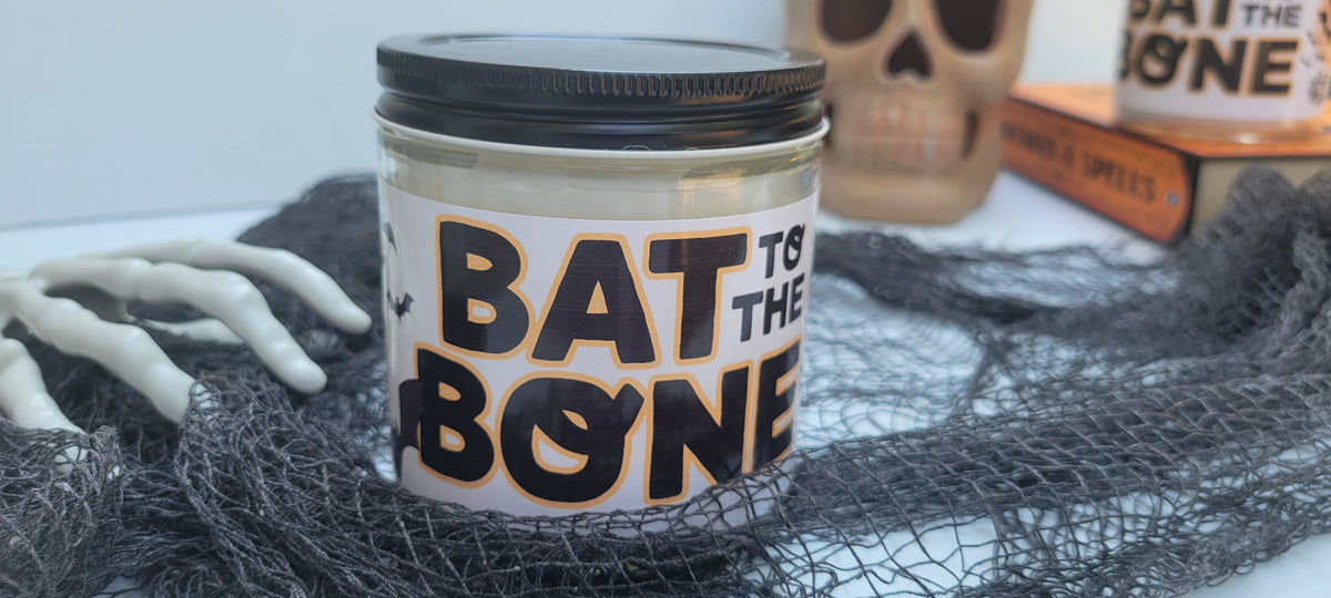 A Halloween-themed candle in a clear glass jar with a black lid, featuring a white label that reads 'BAT TO THE BONE' in black and orange. The candle is styled on a white surface with black spiderwebs, a skeleton hand prop, and a skull in the background.