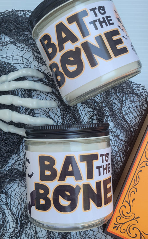 Two Halloween-themed jar candles with black lids and white labels reading 'BAT TO THE BONE' in black and orange lettering, styled with fake skeleton hands, black netting, and an orange decorative piece.