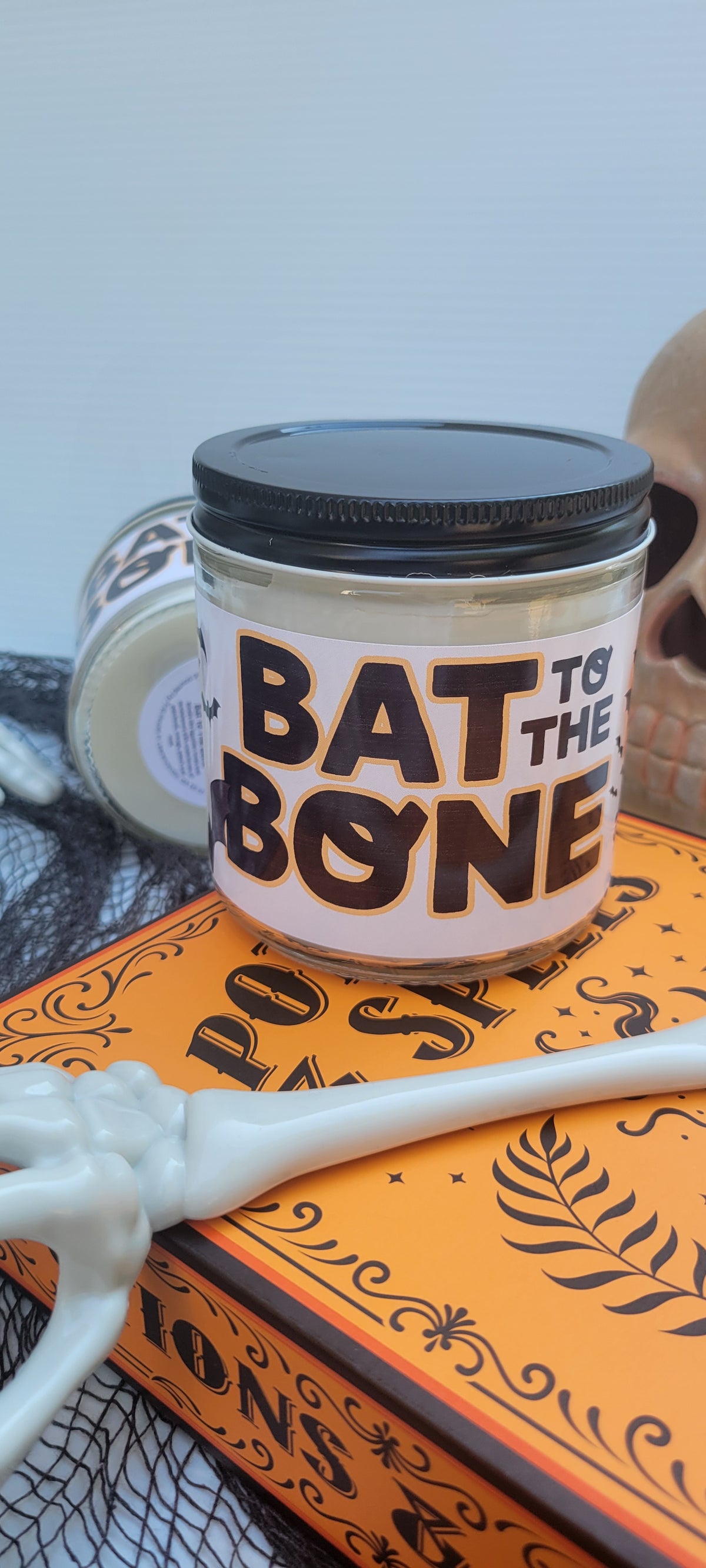 A Halloween-themed jar candle with a black lid and a white label reading 'BAT TO THE BONE' in bold black letters, styled on an orange book with skeleton props.