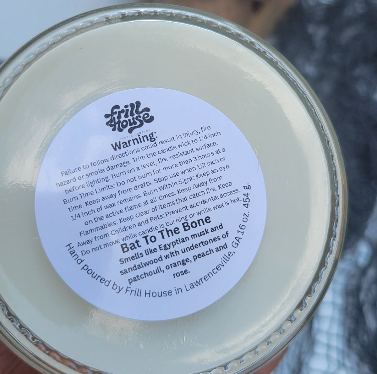 A hand holding a round glass jar of wax candle, showing the bottom label that reads 'Bat To The Bone' by 'The Witch Shop', with a skull decoration in the background.