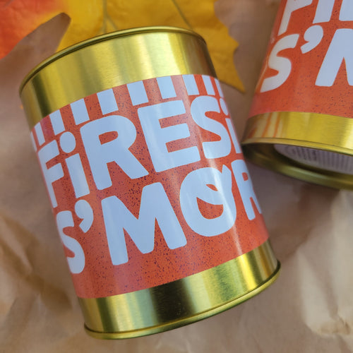 A close-up of a cylindrical tin with a metallic gold top and bottom and a terracotta orange label featuring the text 'FIRE IS S'MORE' in large white letters. Another similar tin is partially visible in the background along with autumn leaves and brown paper.