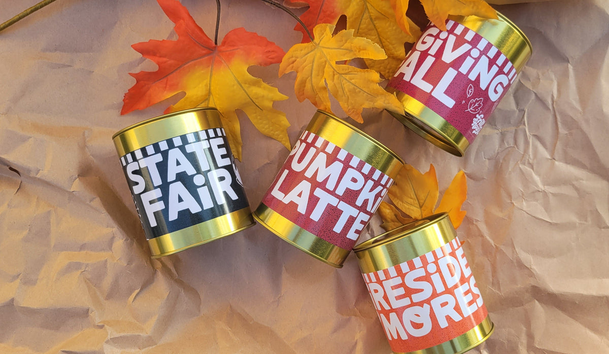 Four cylindrical cans with gold rims and colorful labels arranged on crumpled brown paper with autumn leaves. The labels feature bold white text on red, orange, and navy backgrounds.
