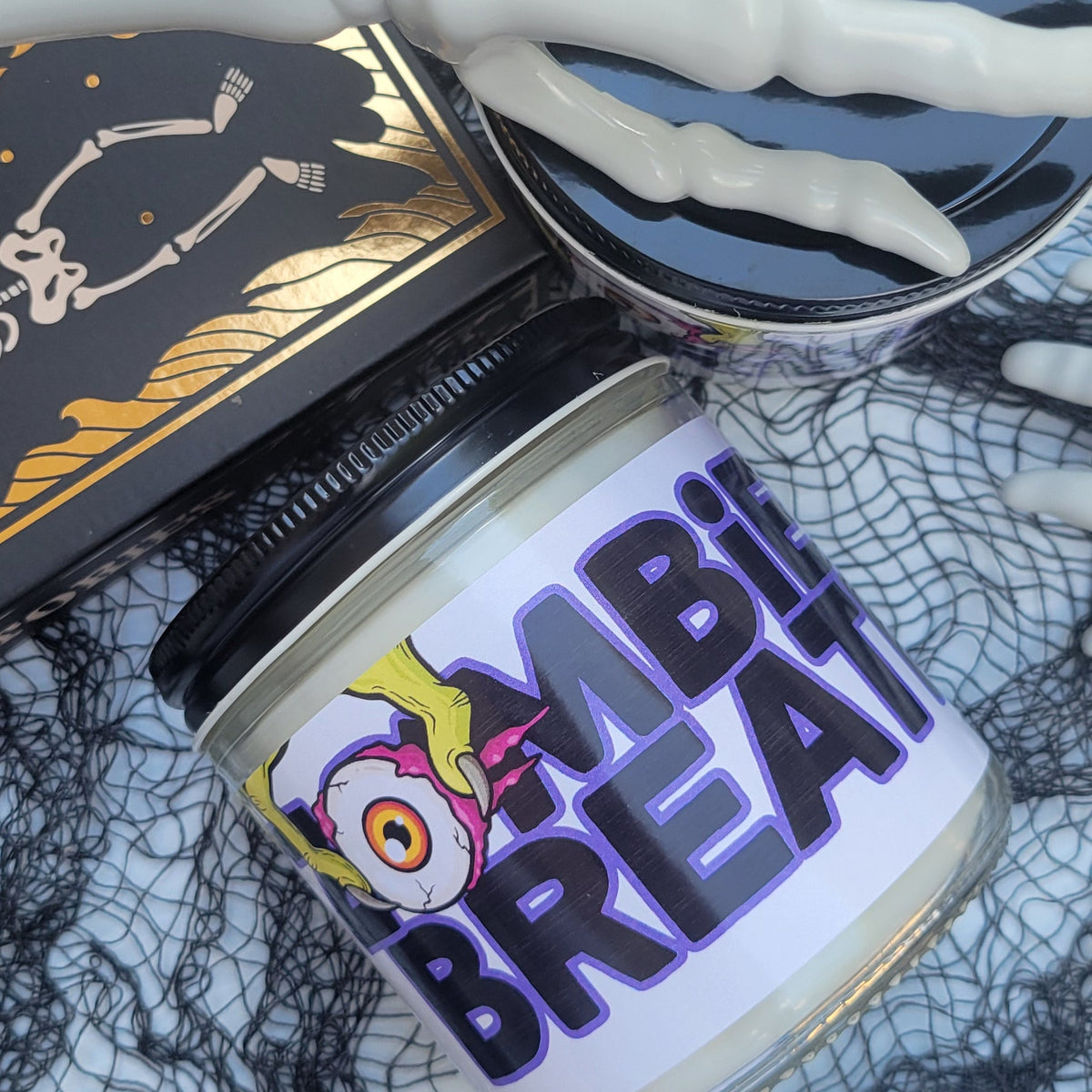 A Halloween-themed candle jar labeled 'ZOMBIE BREATHE' with a black lid, styled with a skeleton hand, spiderweb fabric, a tarot card, and a skull on a white background.