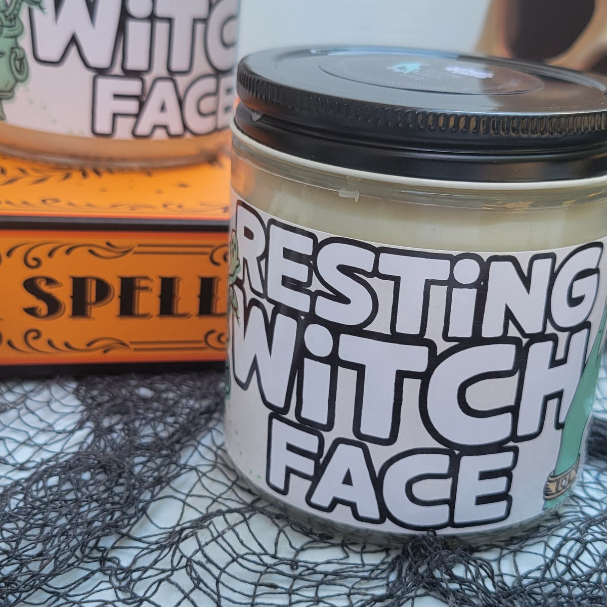 A jar of 'Resting Witch Face' cosmetic product with a black lid and white label, styled on a black webbed fabric with a skull, skeleton hand, and orange book in the background.