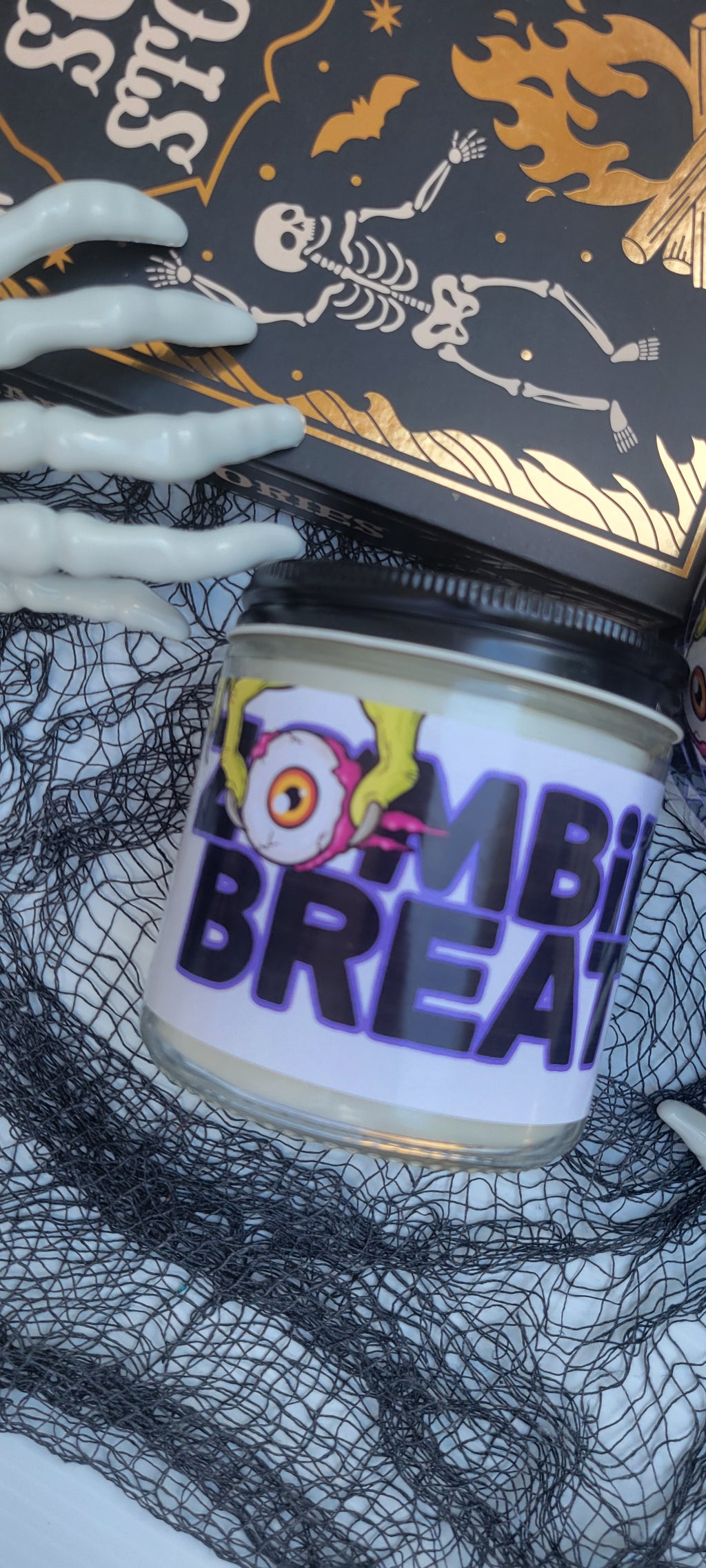 A Halloween-themed jar candle labeled 'Zombie Breath' with a black lid, shown alongside a black box with gold artwork and white skeleton hands on a black net.