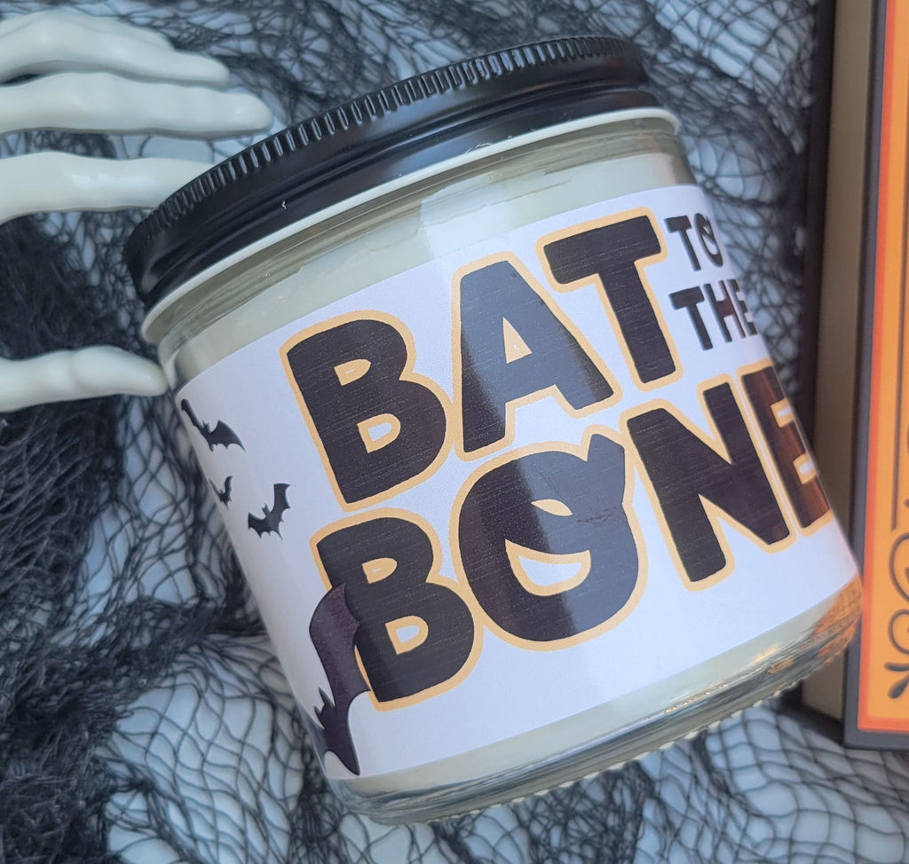 A Halloween-themed jar candle with a black lid and a white label reading 'BAT TO THE BONE' in black letters with orange outlines, styled with black tulle netting and skeleton hands.