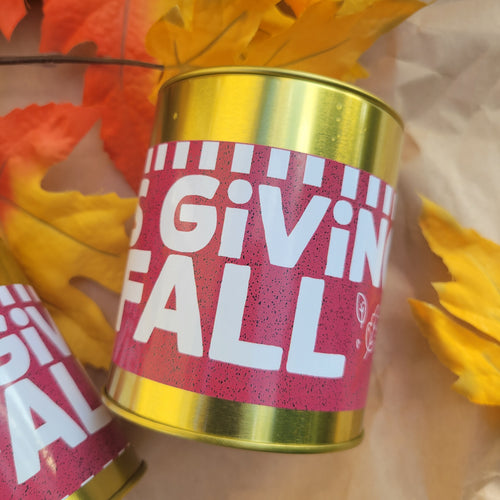 A close-up of a cylindrical tin with a metallic gold top and bottom and a maroon label that reads 'It's Giving Fall' in large white letters. The tin is styled with autumn leaves on a light brown surface.