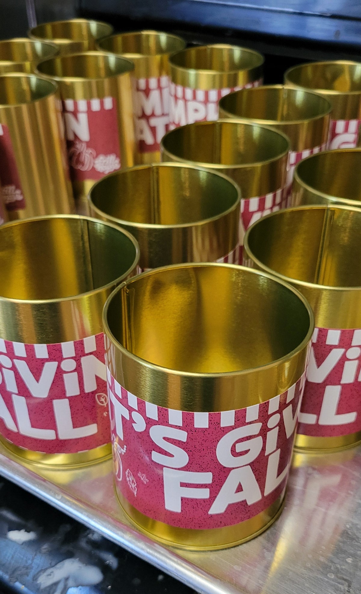 A close-up of several metallic gold cylindrical cups arranged on a metal tray. The front cup has a maroon wrap label with white text reading 'IT'S GIVING FALL' while the tins behind it show similar labels.