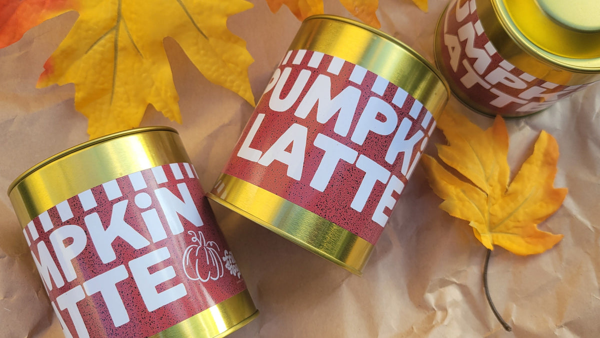 Three gold metal tins of Pumpkin Latte Tea with red labels and white text, arranged on crumpled brown paper with yellow and orange autumn leaves.