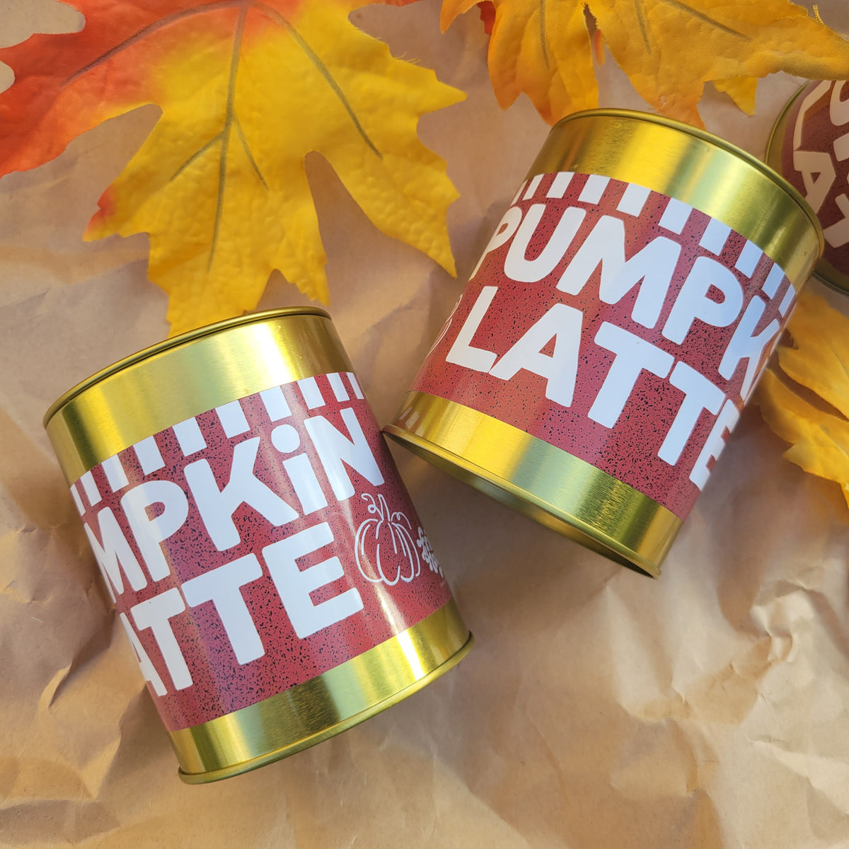 Two cylindrical cans labeled 'PUMPKIN LATTE' with metallic gold rims and pink labels, arranged on brown paper with autumn leaves in the background.