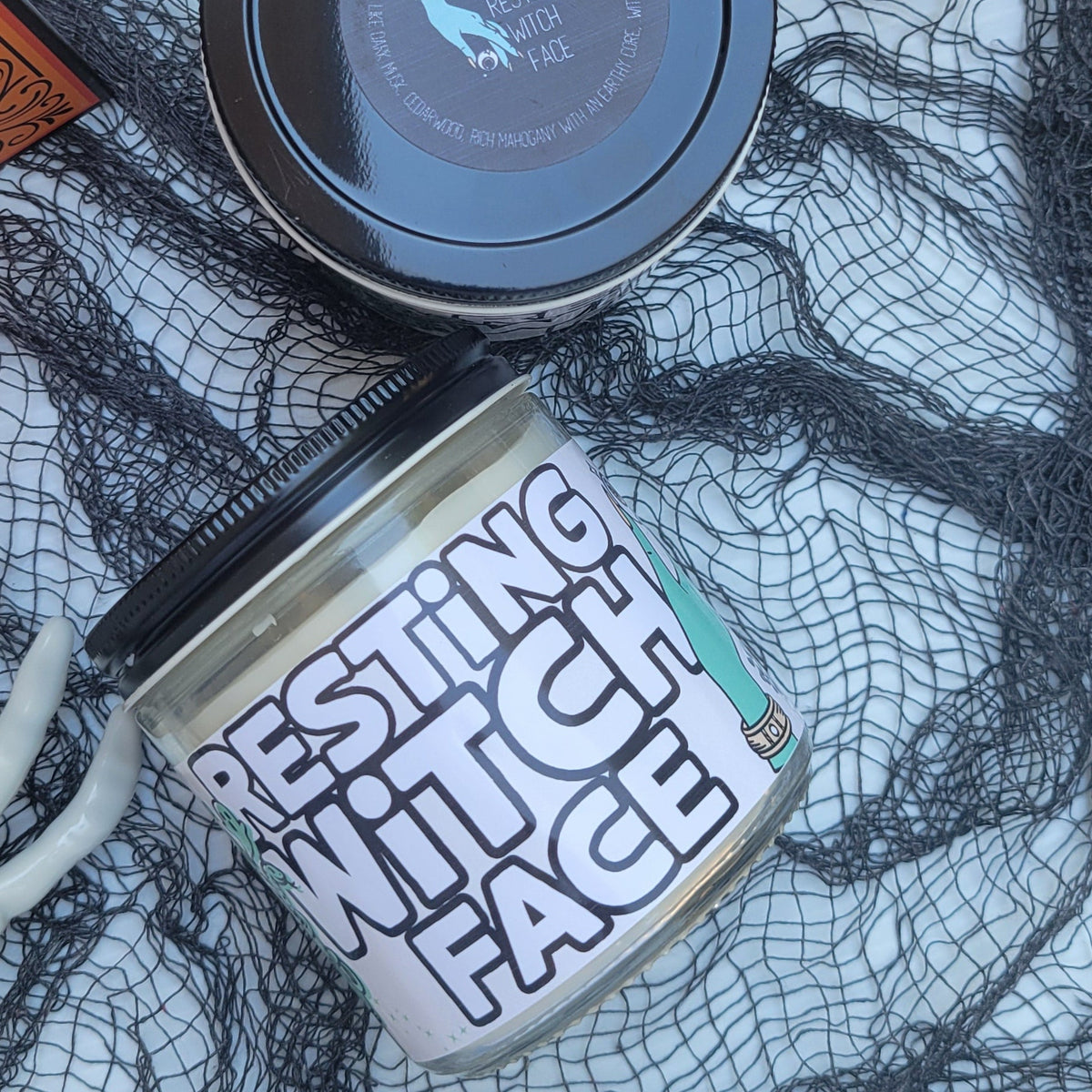 Two cosmetic jars styled on a white surface with black spiderweb fabric and a skull prop. One round jar has a dark lid with a small blue bird logo, and the other cylindrical jar has a white label reading 'TESTING FACE' in black letters.