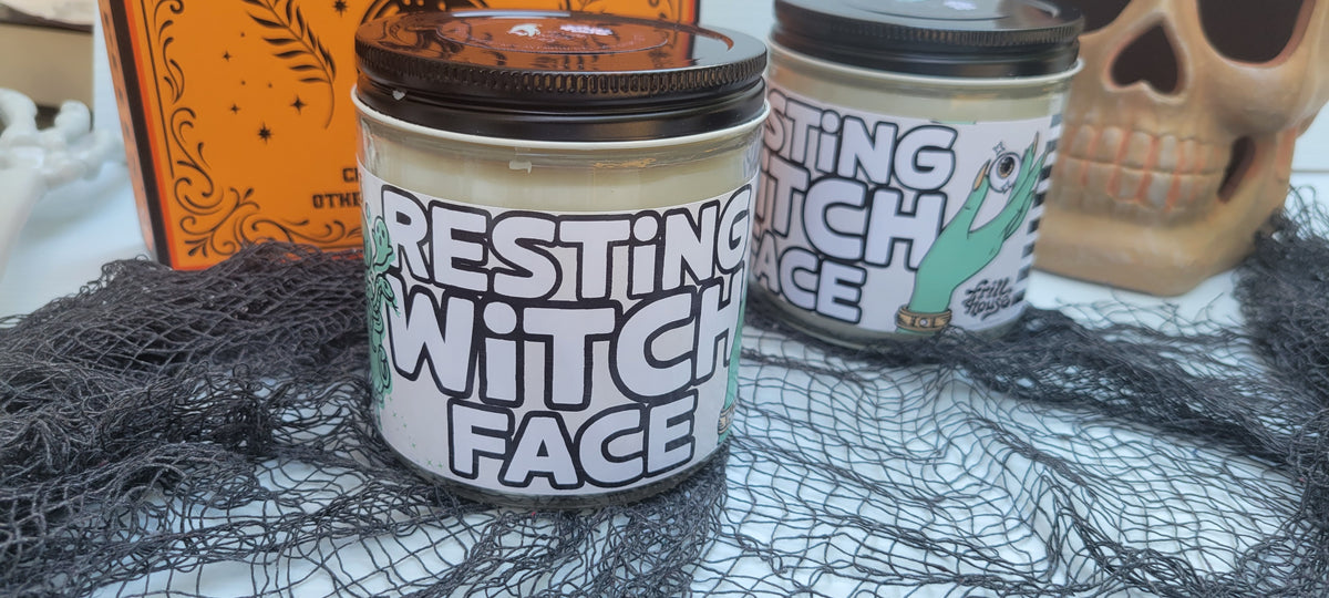 Two jars of 'Resting Witch Face' candle wax with black lids, styled on a white surface with black spiderwebbing, a skull prop, and an orange decorative box in the background.
