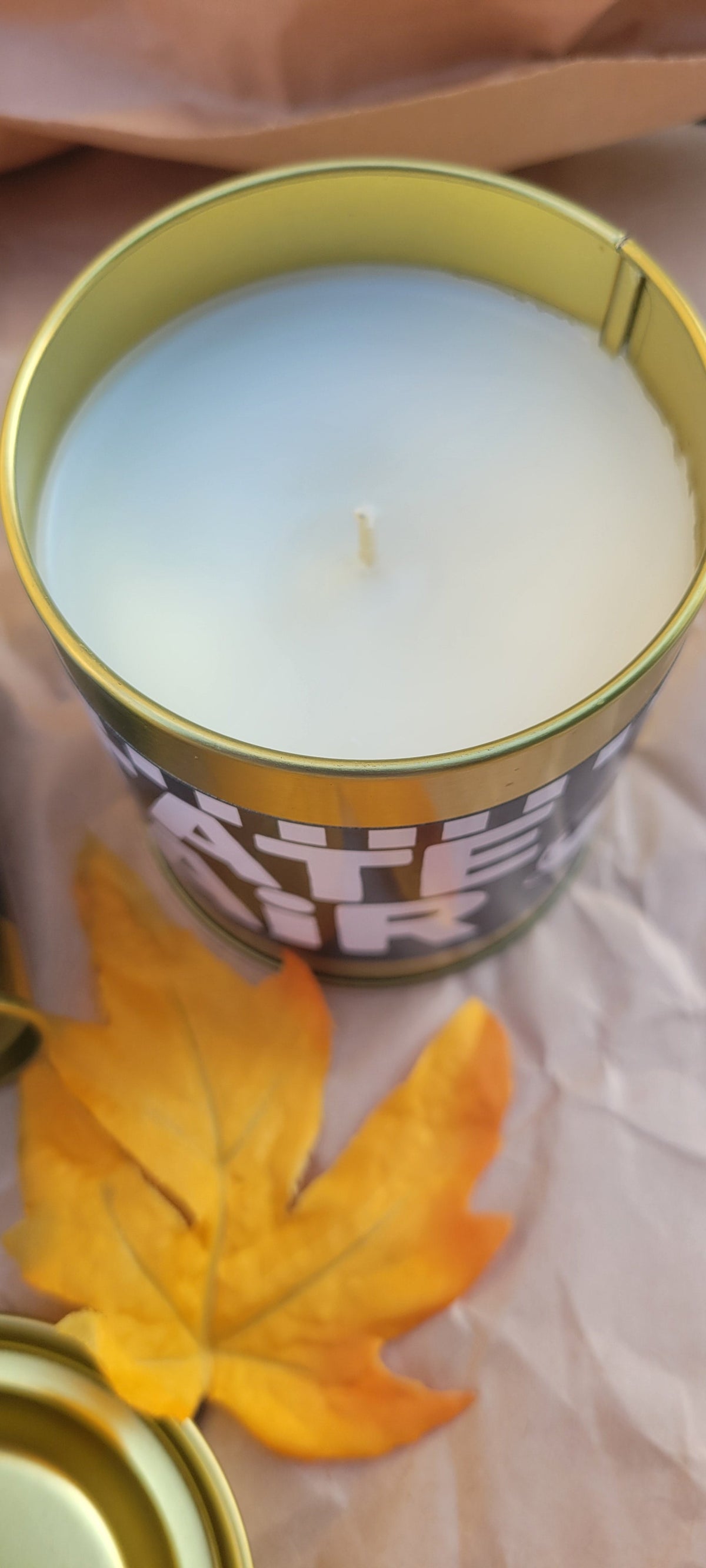 A close-up of a white wax candle in a round metal tin with a gold rim, placed on a light surface next to a yellow-orange leaf. The label on the side of the tin is partially visible.