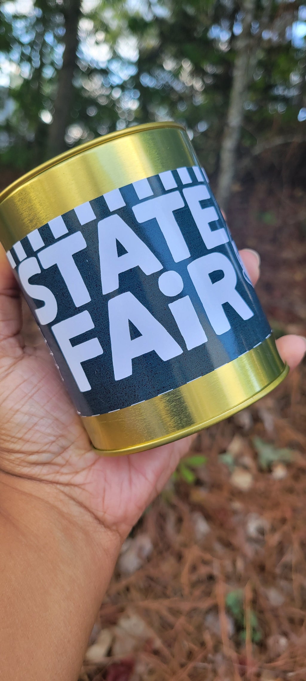 A hand holding a cylindrical tin can with a black label that reads 'STATE FAIR' in large white letters, with metallic gold top and bottom rims, set against an outdoor forest background.
