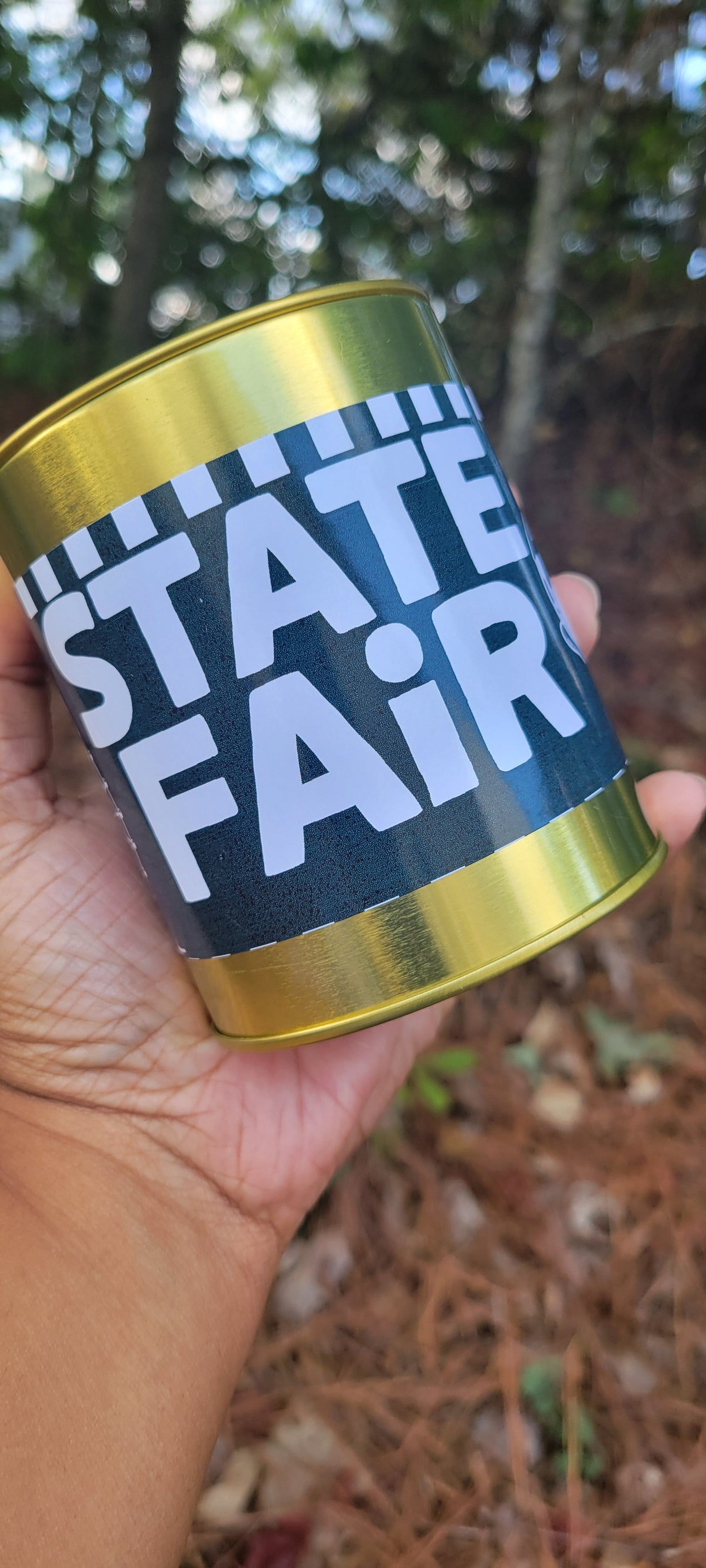 A hand holding a cylindrical tin can with a black label that reads 'STATE FAIR' in large white letters, with metallic gold top and bottom rims, set against an outdoor forest background.