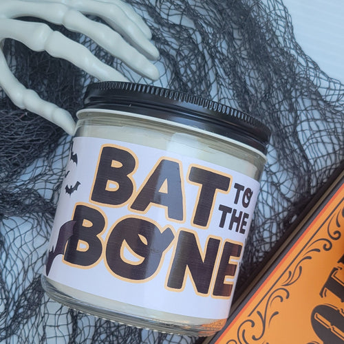 A Halloween-themed jar candle labeled 'BAT TO THE BONE' in black and white lettering, styled with black netting, white skeleton hands, and an orange 'POTATO' sign on a light gray background.