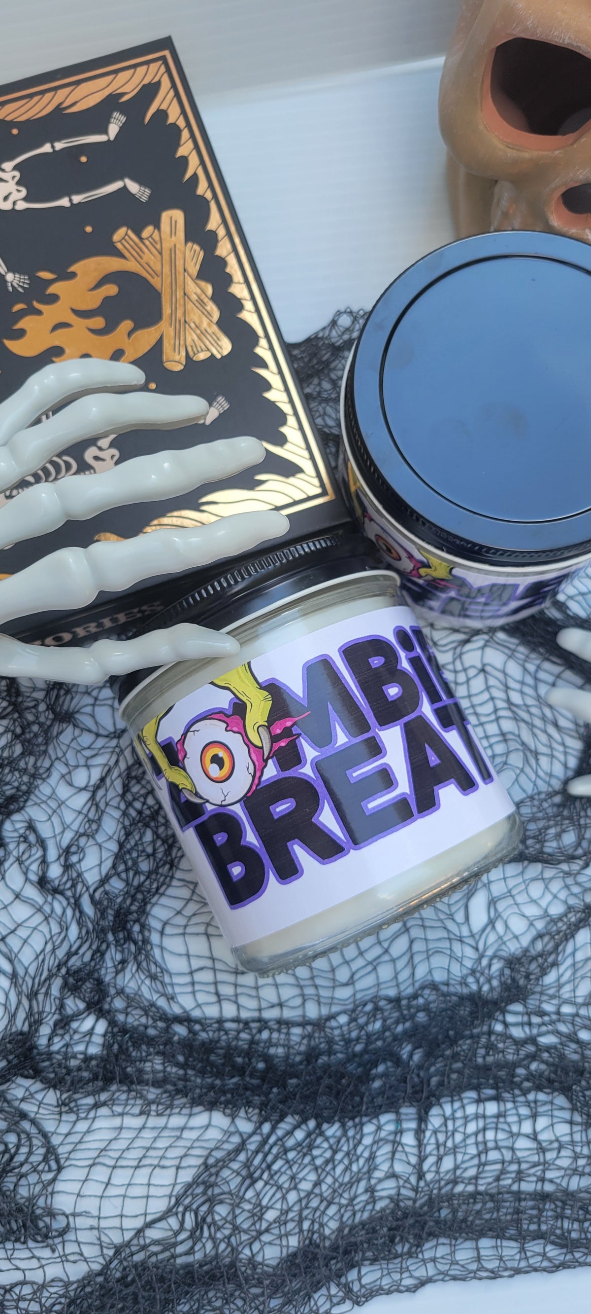A Halloween-themed candle jar labeled 'ZOMBIE BREATHE' with a cartoon zombie face graphic, shown alongside a black box with gold skeleton artwork and a skull prop on a white surface with black netting.