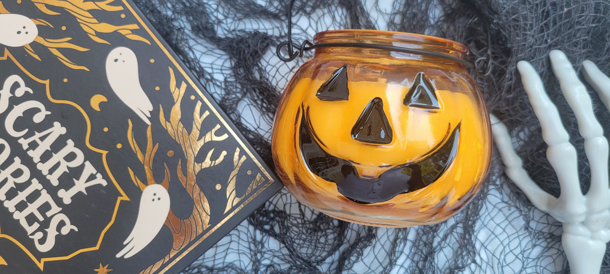 An orange glass pumpkin lantern with a carved jack-o'-lantern face sits on black spiderwebs next to a skeleton hand, with a 'Scary Books' book partially visible in the background.